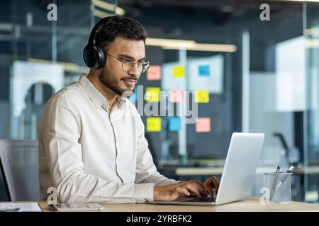 Businessman wearing headphones works diligently on a laptop in a modern office. Surrounded by sticky notes, he embodies focus, technology, and productivity in a contemporary work setting. Stock Photo