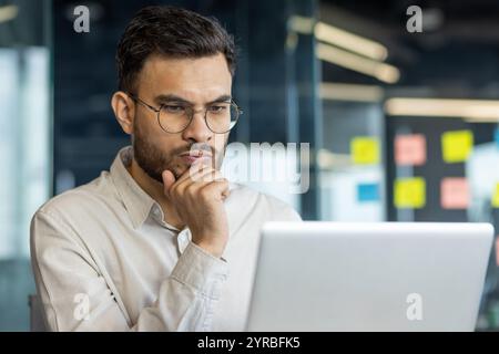 This image features a man, immersed in work, thoughtfully analyzing data on his laptop in an office setting. The professional setting emphasizes focus, concentration, and decision-making aspects. Stock Photo