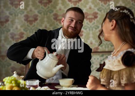 Portrait of classic young gentleman in black tailcoat smiling while pouring tea for young lady sitting at table in vintage setting Stock Photo