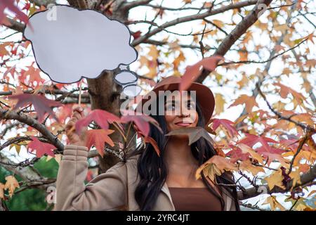 Young latin woman wearing a hat holds an empty thought bubble while posing under a tree with colorful autumn leaves, expressing a sense of contemplati Stock Photo