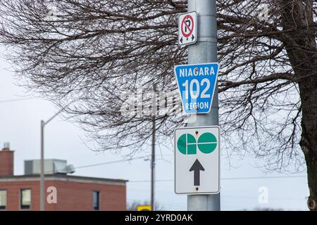 Close-up of a Niagara Region 102 road sign and GO Transit directional sign on a pole, surrounded by urban scenery and trees. Stock Photo