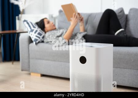 Woman is reading a book on a couch. A white air purifier is on the floor next to the couch Stock Photo