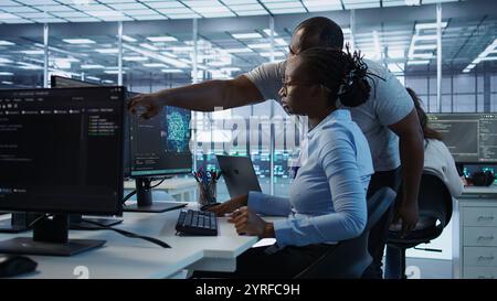 Manager coming to technician desk to give instructions on doing maintenance on artificial intelligence neural networks. Supervisor overseeing worker using AI system software, camera B Stock Photo