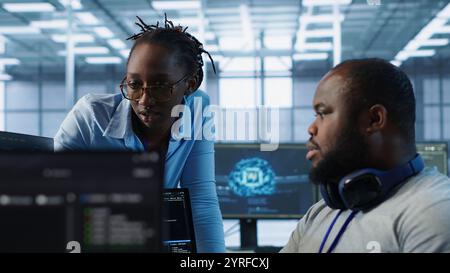 Engineer working with colleague on computer in high tech data center establishment monitoring hardware energy consumption. Team of technicians using PC to check for system failures, camera A Stock Photo