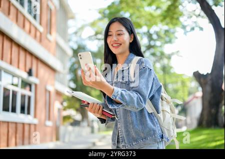 A beautiful, happy Asian female college student in a denim jacket, carrying her backpack and books, enjoys a video call with her friends through her s Stock Photo