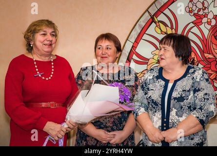 Three Russian friends, aged 60 and 70, hold a bouquet of flowers and congratulate their friend on a special occasion. They are looking sideways, dress Stock Photo