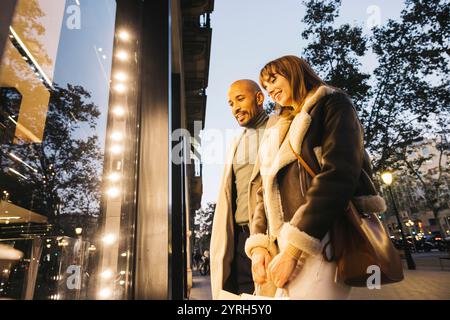 A cheerful couple looking at a window shop in the city, exploring stores on a bright day. Stock Photo