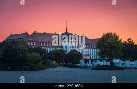 The grand hotel at sopot beach stands majestically against a vibrant sunset sky, with closed beach umbrellas and lush greenery in the foreground, crea Stock Photo