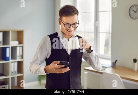 Man standing with a cup coffee at office and reading messages on his mobile phone. Stock Photo