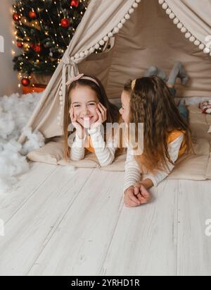 Two happy sisters share a magical Christmas moment inside a cozy teepee. Their bright smiles and comfortable poses capture the joy of the season. The Stock Photo