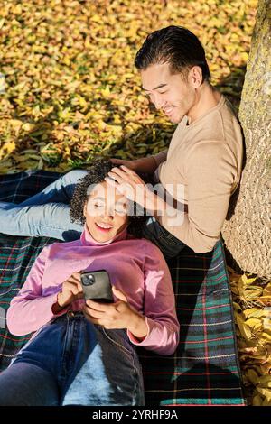 A smiling couple relaxes on a blanket amidst fallen autumn leaves The woman looks content while using her smartphone, as the man tenderly touches her Stock Photo