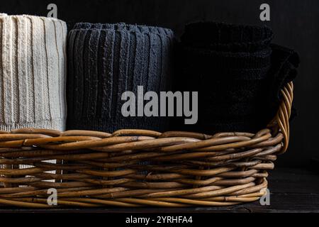 Soft, textured knitted fabrics in black, gray, and cream are neatly rolled, displayed in a rustic wicker basket against a dark wooden background, evok Stock Photo
