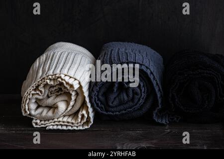 Three neatly rolled knitted sweaters in cream, gray, and black showcased on a dark background The warm textures and neutral shades highlight a cozy, m Stock Photo