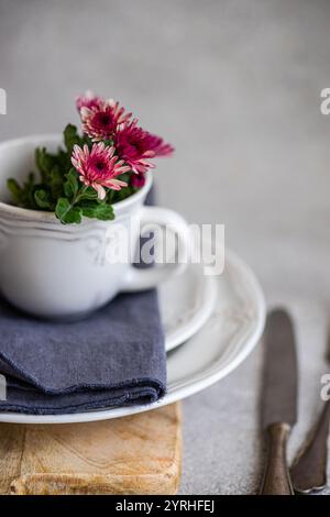 A rustic white ceramic cup filled with vibrant pink flowers sits on a wooden surface A soft gray napkin lies beneath, creating a cozy, inviting atmosp Stock Photo
