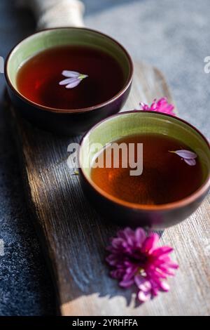 Two ceramic cups filled with tea sit on a textured wooden board Each cup has floating pink flower petals, accented by nearby pink blooms, creating a s Stock Photo