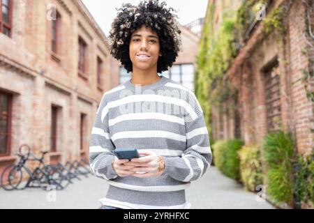 A mixed race woman with vitiligo enjoys a relaxing day on her university campus She stands outdoors, holding her phone with a joyful expression Brick Stock Photo