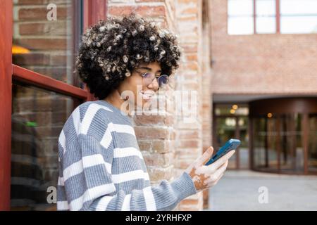 A mixed race woman with vitiligo smiles while using her smartphone, enjoying a relaxing day at the university Brick walls and large windows create an Stock Photo