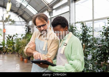 Two garden store employees in aprons discuss work while using a digital tablet in a greenhouse filled with potted plants They focus on teamwork and te Stock Photo
