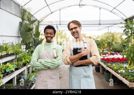 Two smiling employees in aprons stand inside a bustling garden center, surrounded by vibrant plants and flowers The cheerful atmosphere reflects passi Stock Photo