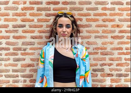Stylish young woman stands confidently in front of a textured brick wall. She wears a black top, colorful jacket, and sunglasses on her head. Stock Photo