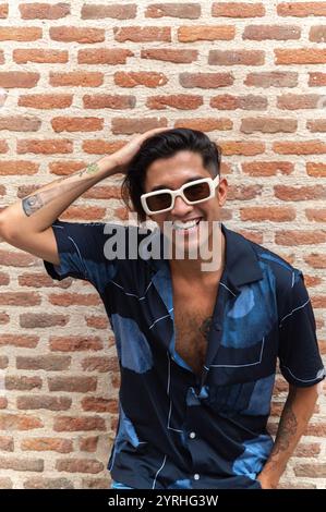 A joyful young man with slicked-back hair and fashionable white sunglasses stands before a red brick wall, laughing as he touches his hair. Stock Photo