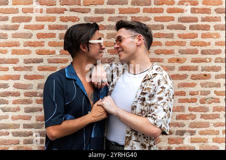 Two young men share a tender moment together, standing close and gazing at each other fondly against a textured brick wall background. Stock Photo