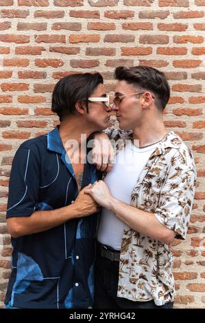 A heartfelt moment between two stylish men as they share a kiss and hold each other tightly in front of a rustic brick wall. Stock Photo