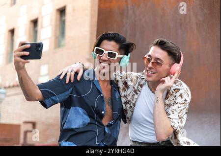 Two fashionable young men pose for a selfie, one wearing sunglasses and headphones, the other with earphones, both sharing a joyful moment on a sunny Stock Photo