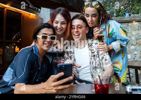 A diverse group of four friends sharing a joyful moment while taking a selfie at a cozy outdoor bar, with drinks in hand during a lively evening. Stock Photo
