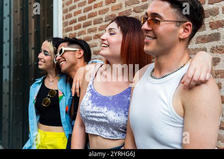 Four young adults stand together, laughing and enjoying themselves outside against a red brick wall. They are dressed in fashionable summer attire Stock Photo