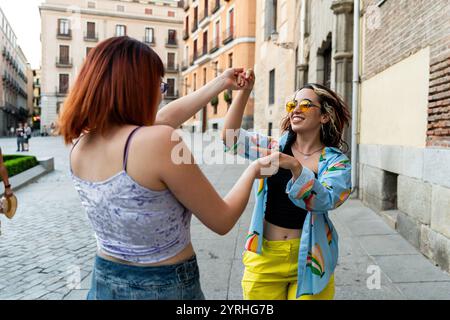 Two happy young women dancing and laughing together on a city street, showcasing joy and friendship in a casual urban setting. Stock Photo