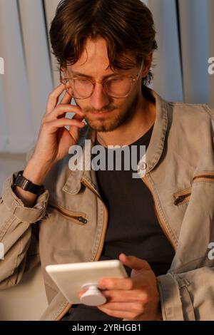 A young man with glasses and casual attire, deep in concentration while using a digital tablet indoors He exudes a relaxed yet focused vibe, perfect f Stock Photo