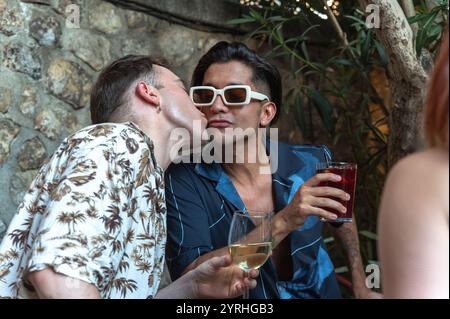 A tender scene captures one man kissing another on the cheek while holding glasses of wine at a lively garden party, reflecting a moment of love and c Stock Photo