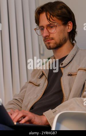 A man focused on his laptop while sitting, wearing a casual jacket and glasses He appears concentrated in a relaxed, indoor setting, reflecting modern Stock Photo