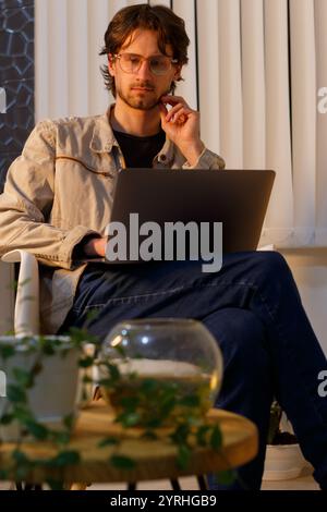 Man in casual attire works on a laptop in a warmly lit room He appears thoughtful, possibly contemplating ideas or solutions Surrounding plants add a Stock Photo
