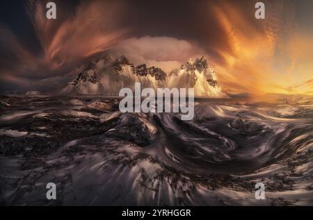 A breathtaking view of snow-covered mountains under a dramatic sky at sunset. The fiery hues and swirling clouds create a mesmerizing and otherworldly Stock Photo