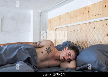 Young man with tattoos lying on his side in bed with a thoughtful expression. The room has wooden elements and gray bedding, creating a cozy, relaxed Stock Photo