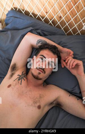 A relaxed man with tattoos lies on a bed looking at the camera. The cozy setting and casual pose convey a sense of ease and calmness, highlighting the Stock Photo