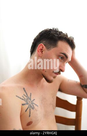 A young man with short hair and tattoos reflects while seated on a wooden chair. The minimalist white background highlights his contemplative expressi Stock Photo