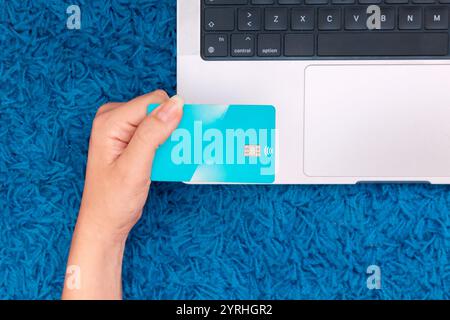 A top view of a person holding a blue credit card while using a laptop on a vibrant blue carpet, representing the ease and comfort of online shopping Stock Photo