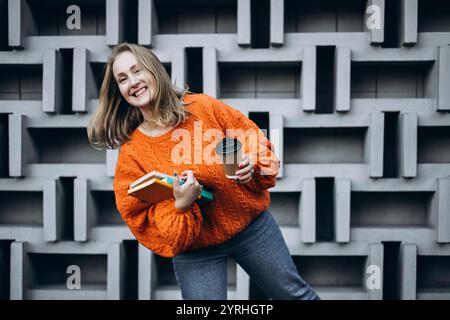 Smiling female student in an orange sweater holds books and a coffee cup while standing against a geometric-patterned wall Casual, modern, and stylish Stock Photo