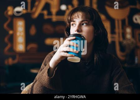 A young woman sips from a blue cup, gazing thoughtfully in a cozy cafe. The warm light and modern decor create a relaxed, inviting atmosphere for cont Stock Photo