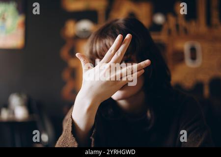 A person sits in a cozy, warmly lit room with brown tones, partially hiding their face behind a casually raised hand. The setting suggests comfort and Stock Photo