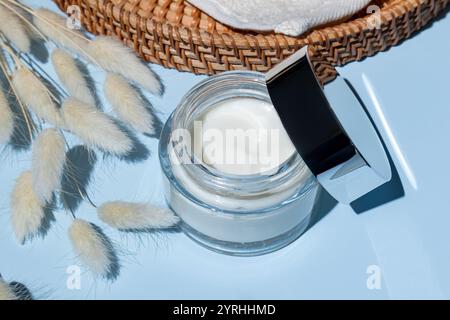 A clear cosmetic jar filled with creamy skincare product is arranged beside soft, white pampas grass on a light blue surface Natural and luxurious bea Stock Photo