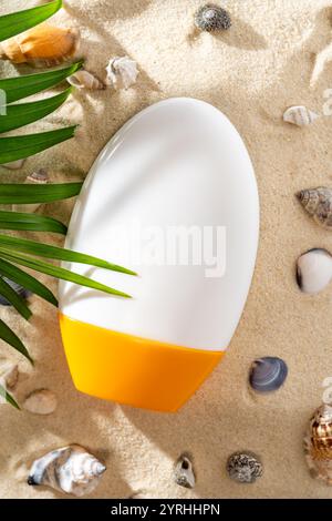 A close-up of a sleek sun protection cream bottle with an orange cap resting on sandy beach surroundings, decorated with seashells and a palm leaf, ca Stock Photo