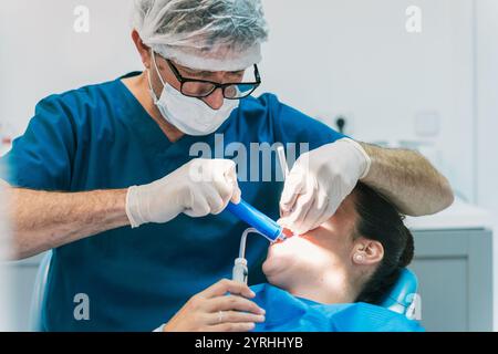 A dentist focuses intently while performing a dental procedure on a patient in a modern clinic. The dentist wears a mask, gloves, and a protective cap Stock Photo