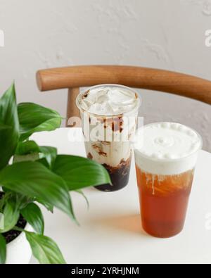 Two iced beverages, creamy coffee and frothy tea, rest on a round white table beside a lush green plant A wooden chair enhances the cozy, inviting set Stock Photo