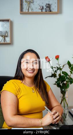 Smiling young woman in a yellow top sits confidently at a desk in an office setting Behind her, a vase with red roses adds a touch of elegance to the Stock Photo