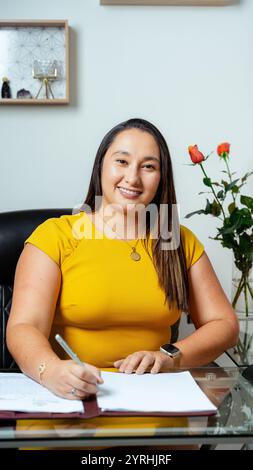 A confident woman in a yellow dress smiles while signing documents at a desk in a modern office setting, with roses in the background Ideal for busine Stock Photo