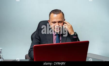 A businessman in a black suit converses on his smartphone, with a red laptop open in front The setting portrays a professional and modern office envir Stock Photo
